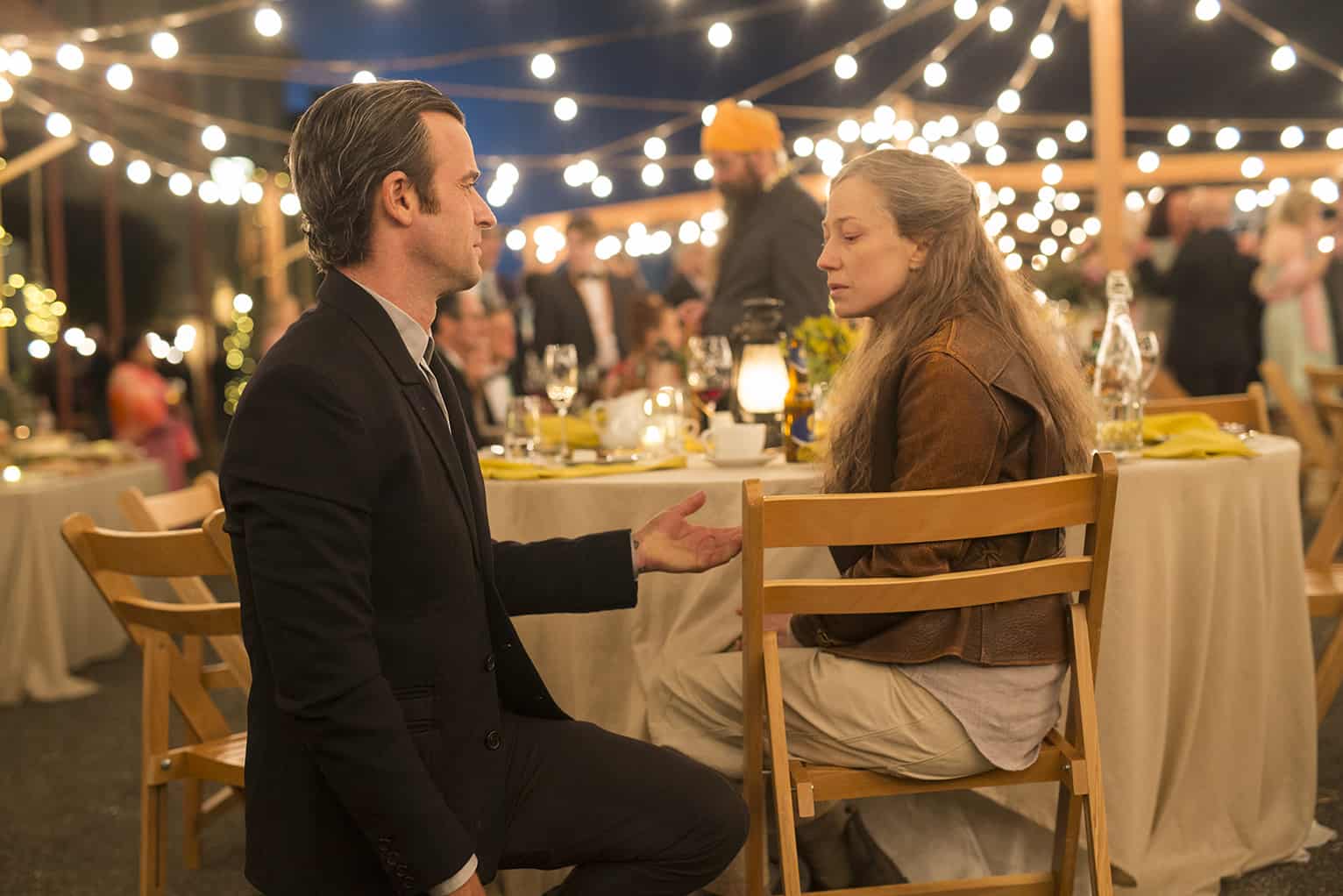 A man kneels in front of a woman at an outdoor table in this image from Warner Bros. Television.