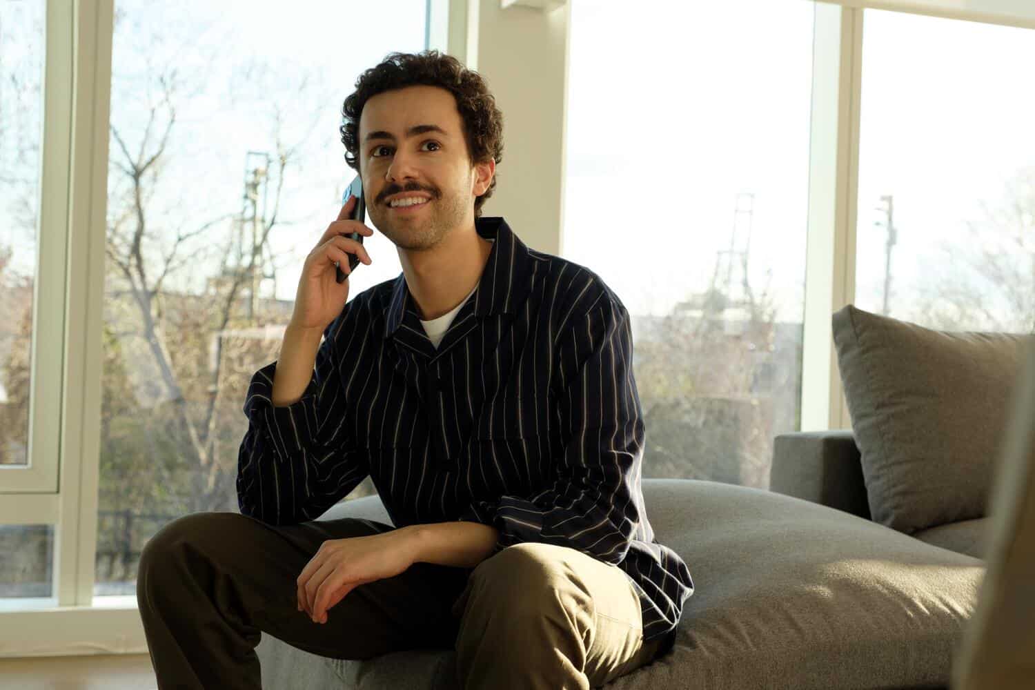 A man sits on the couch and talks on a smartphone in this image from A24 Television.