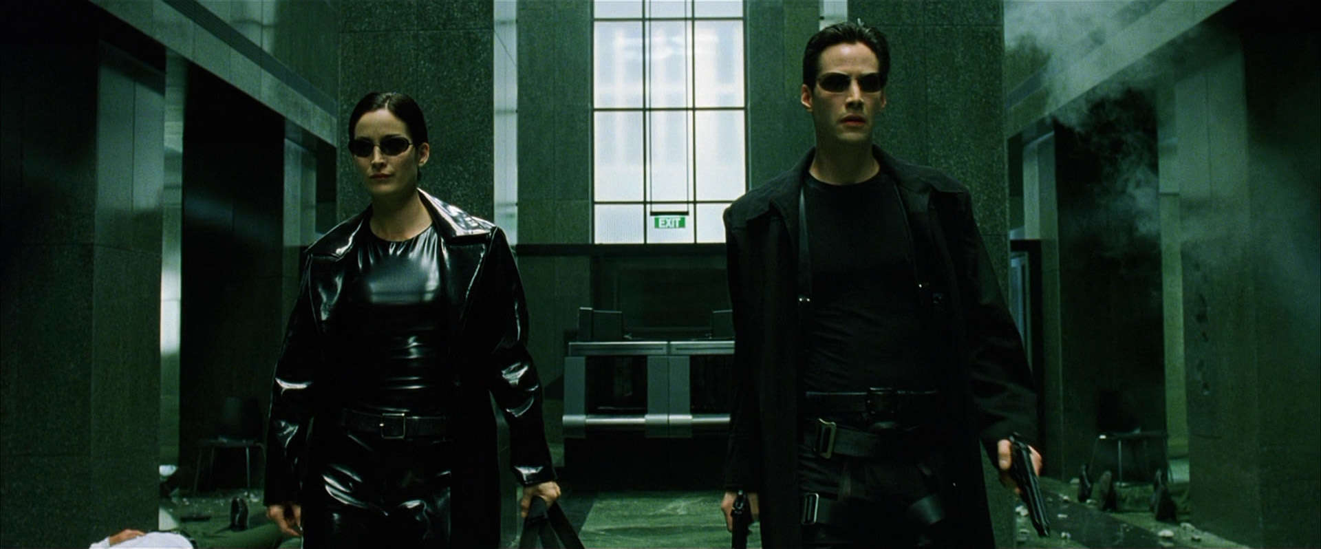 A woman and a man wearing sunglasses hold guns and walk down a hall in this image from Warner Bros. Pictures.