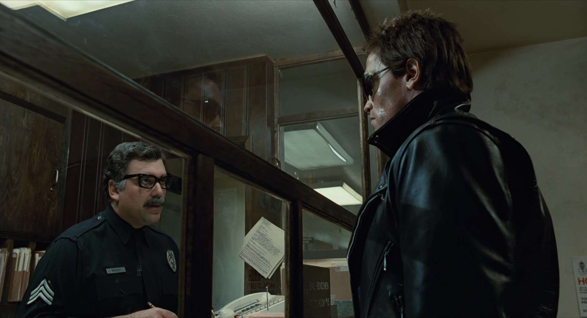 A man in sunglasses speaks with an officer behind a glass wall in this image from Cinema '84.