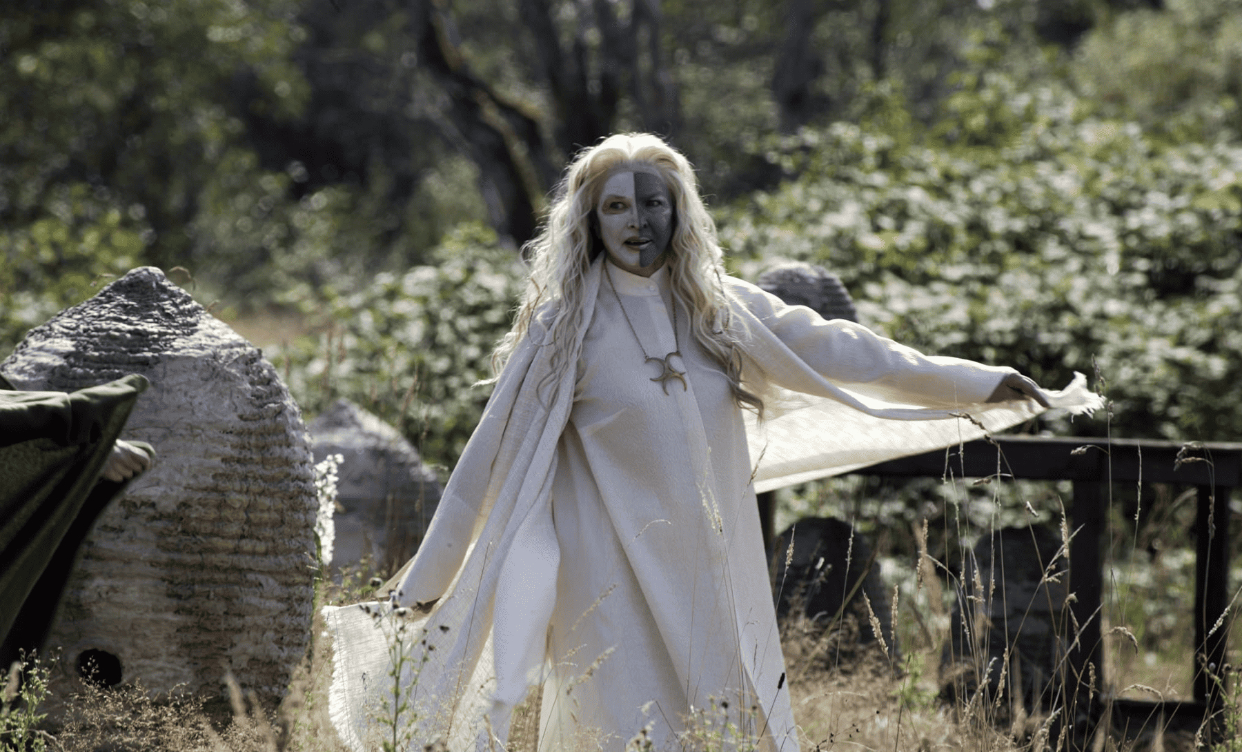 A neo-pagan woman with her face half-painted blue and dressed in a white, flowy dress seems to walk airplane-style across a field in this image from Warner Bros.