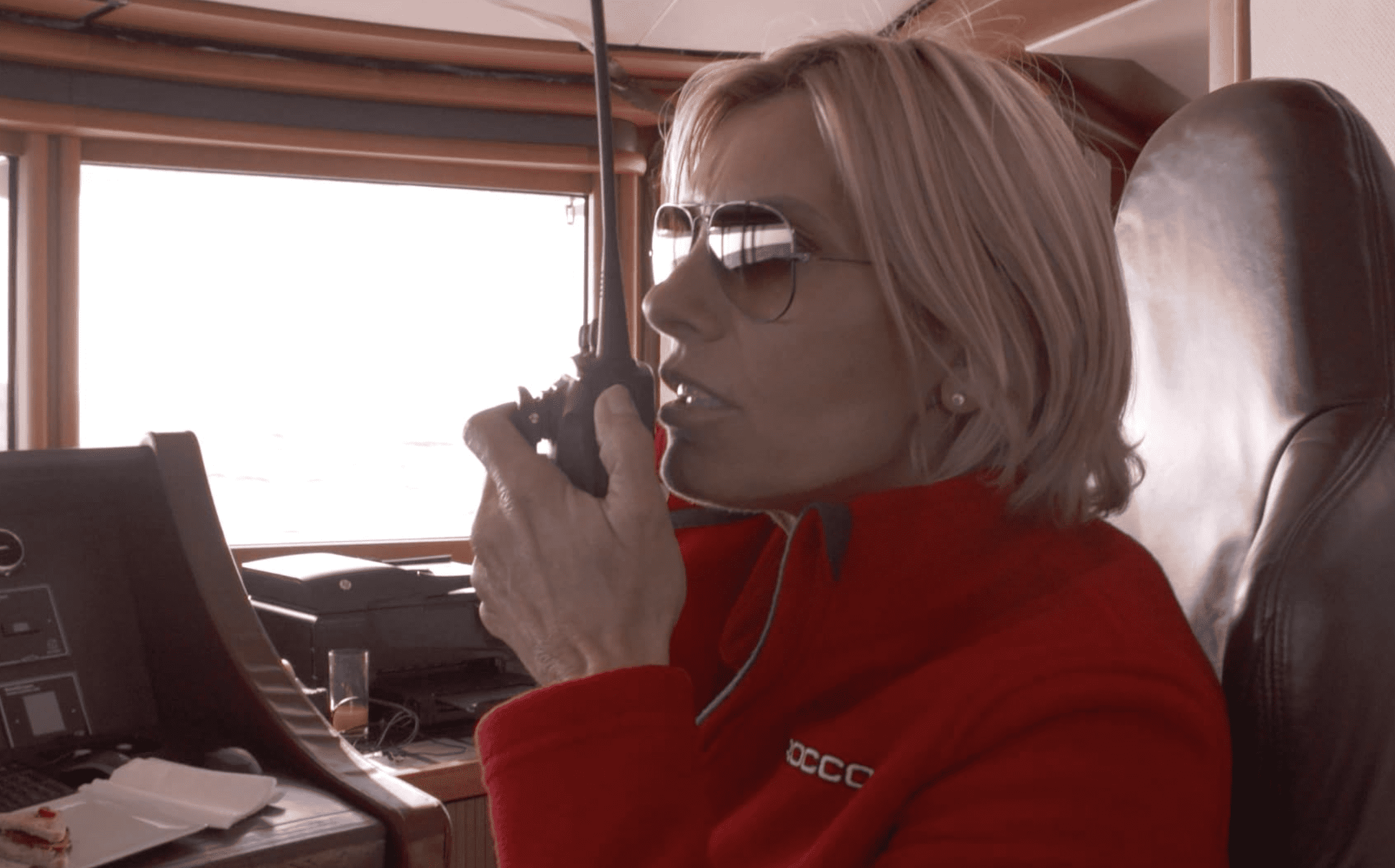 A woman with blond hair, sunglasses, and a red jacket uses a radio from the wheelhouse in this image from 51 Minds Entertainment.