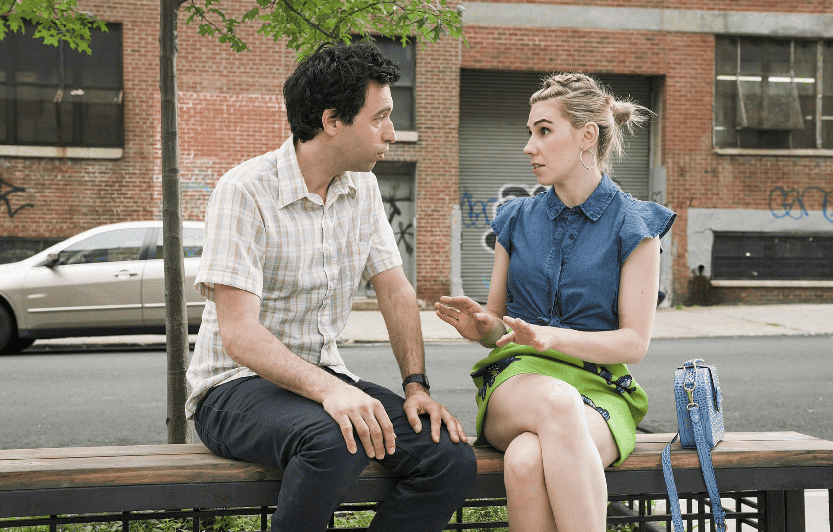 A man and a woman have a conversation while sitting on a sidewalk bench in this image from Apatow Productions.