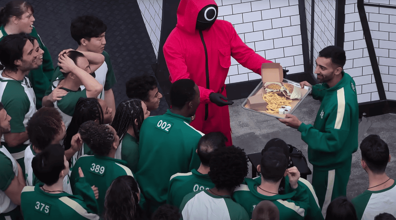 A large group of people in green tracksuits gather around a hooded figure, handing over just one tray of food in this image by Studio Lambert.