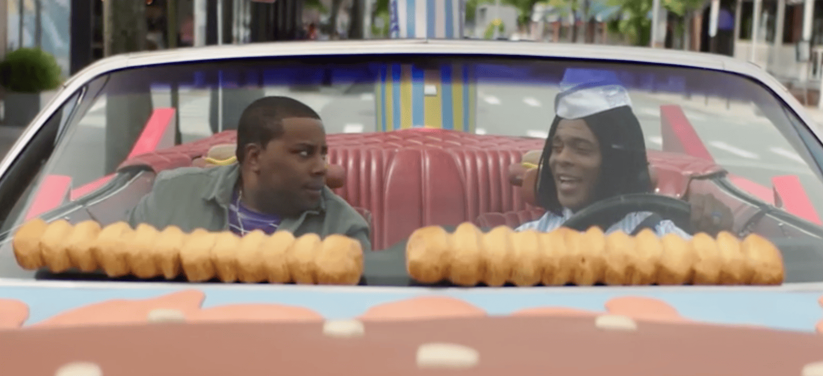 Two men sitting inside a convertible car with the hood off and with what appear to be hamburger buns lining the windshield in this image from Paramount Pictures.