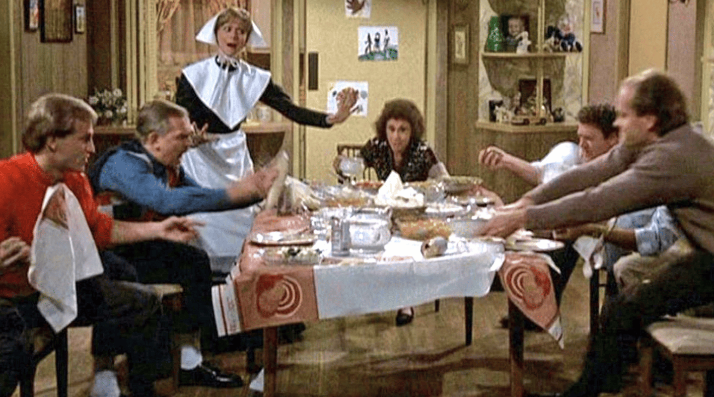 A group of adults gets rowdy around a Thanksgiving table with a woman standing dressed as a pilgrim in this photo from Charles/Burrows/Charles Productions. 