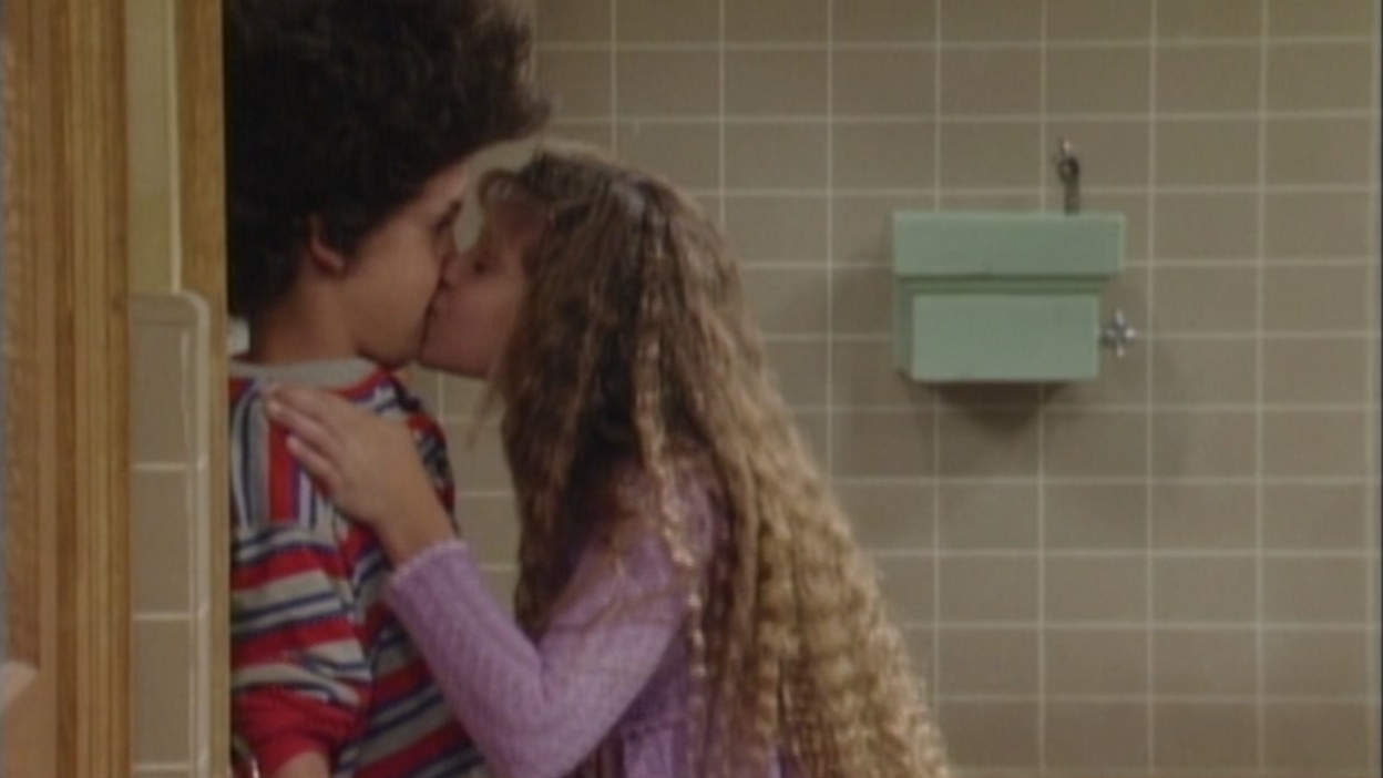 Two middle-school-aged kids share a kiss in the hallway of a school in this image from Touchstone Television.