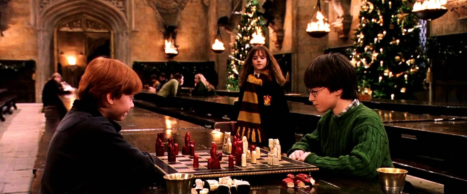 Two boys play chess with a girl looking on and Christmas trees in the background in this image from Warner Bros.