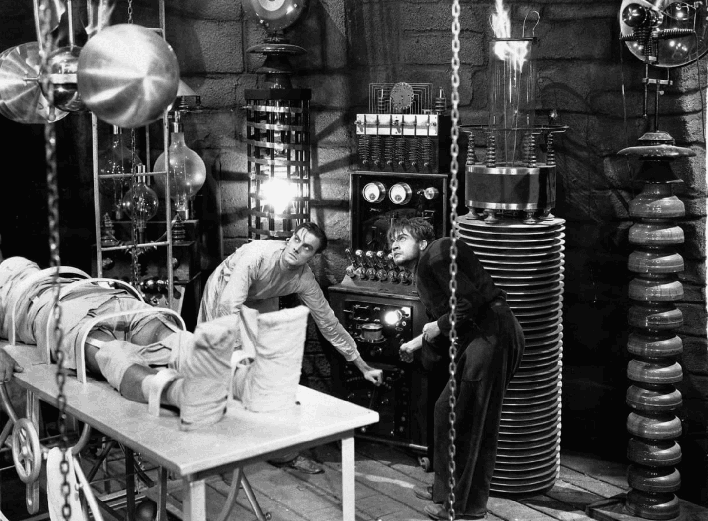 A scientist and his hunchback assistant mess with equipment in a laboratory with a wrapped-up corpse on a table beside them in this image by Universal Pictures.