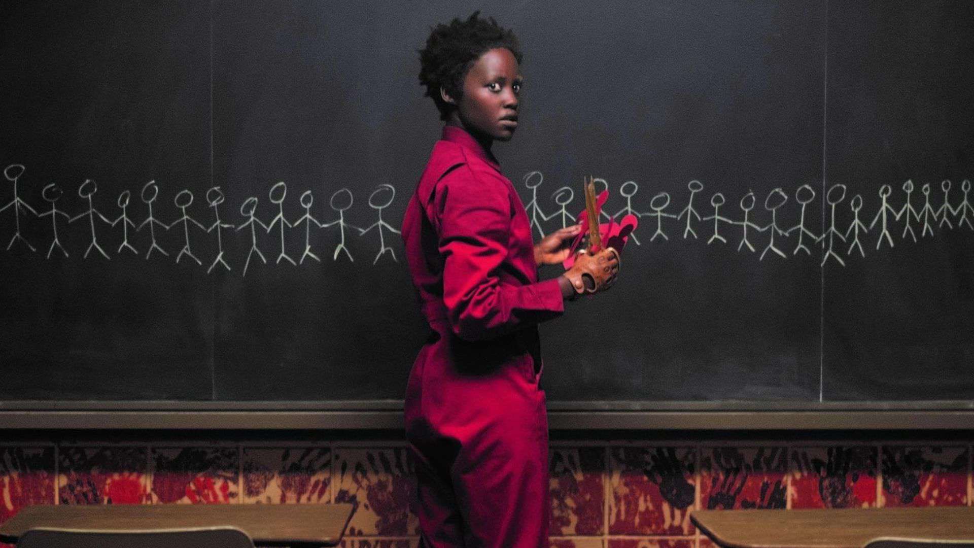 A woman in a red jumpsuit holding gold scissors in front of a chalkboard in this image from Monkeypaw Productions