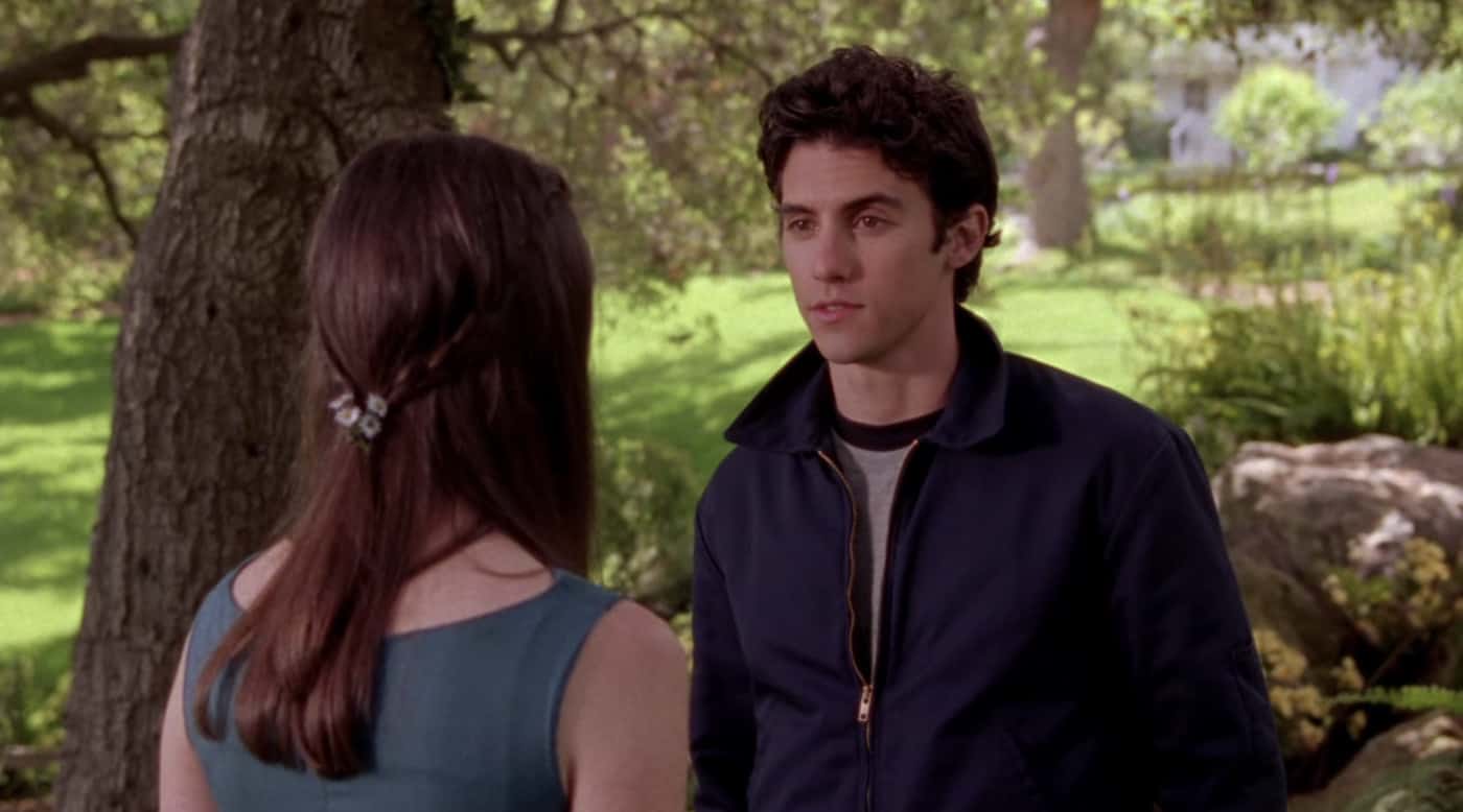 A teenage boy stands in a park with a teenage girl in this image from Warner Bros. Television.
