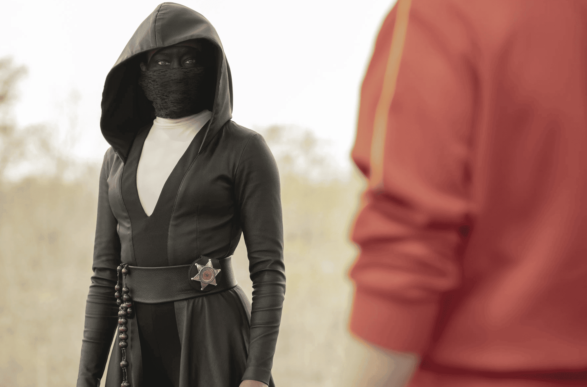 A masked woman with a badge on her hip stands in front of a person in this image from White Rabbit.