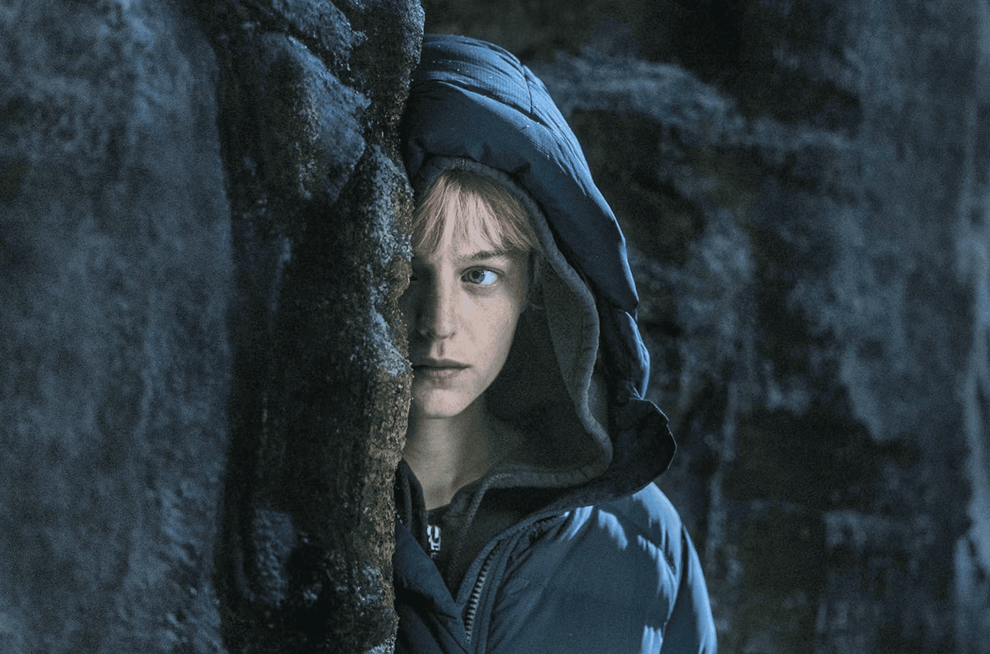 A woman wearing a coat peers from behind a rocky cliff in this image from Mysterium Valley.