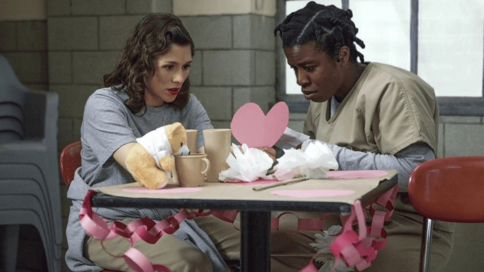 Two women in prison uniforms sit at a table with Valentine’s Day decorations in this image from Tilted Productions.
