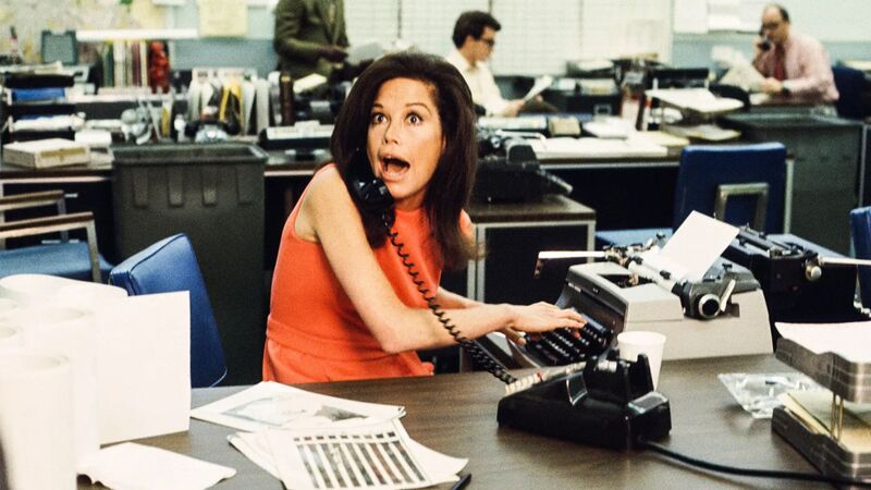A woman in an orange dress talks on the phone while typing on a typewriter in this image from MTM Enterprises.