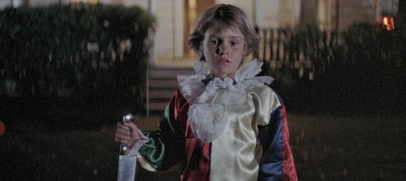 A young boy in a clown costume stands outside holding a knife in this image from Compass International Pictures.