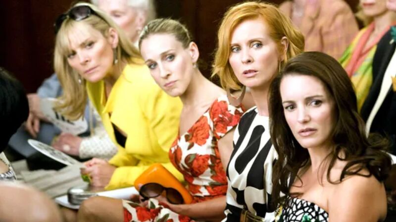 Four women sitting next to each other all looking at something in this image from Darren Star Productions.