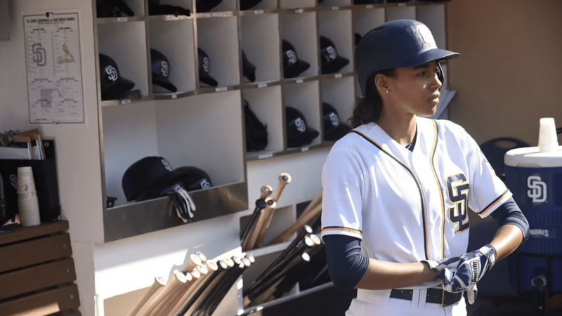 A female baseball player dressed in uniform removes her batting gloves in the dugout in this image from Barnstorm Films.