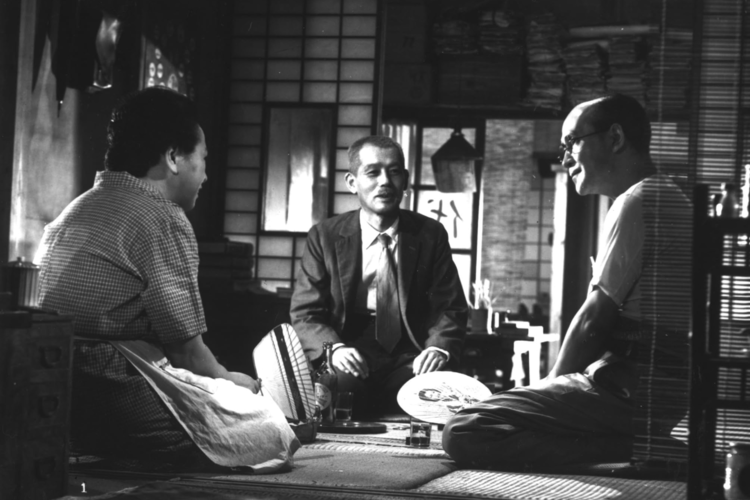 A monochrome image of three individuals engaged in a conversation, seated on tatami mats in a traditional wooden house in this image from Shochiku.