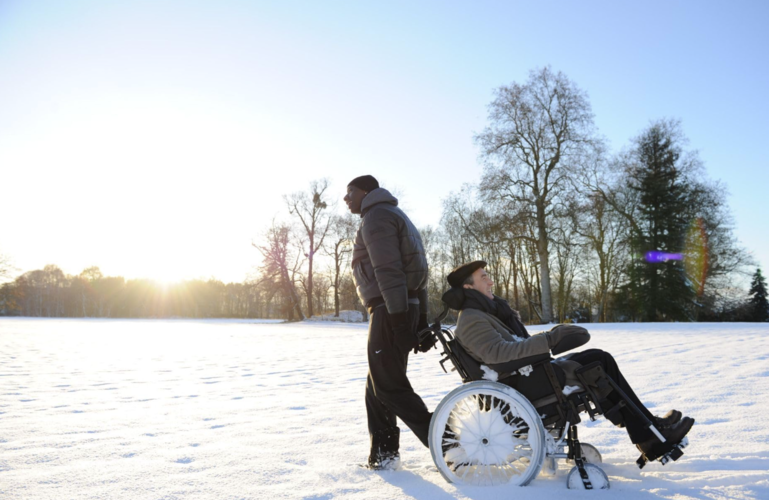 In a calm, snow-covered landscape, a man pulls another man in a wheelchair backward in this image from Quad Productions.