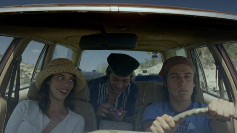 Three friends share a cheerful ride in a vintage car, with the woman glancing at the driver in this image from Anhelo Producciones.