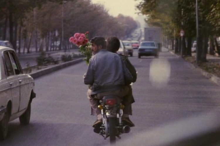 Two people ride a motorcycle down a tree-lined street, with one holding a bright bouquet of red flowers, in an urban setting in this image from Kanun Parvaresh Fekri Productions.