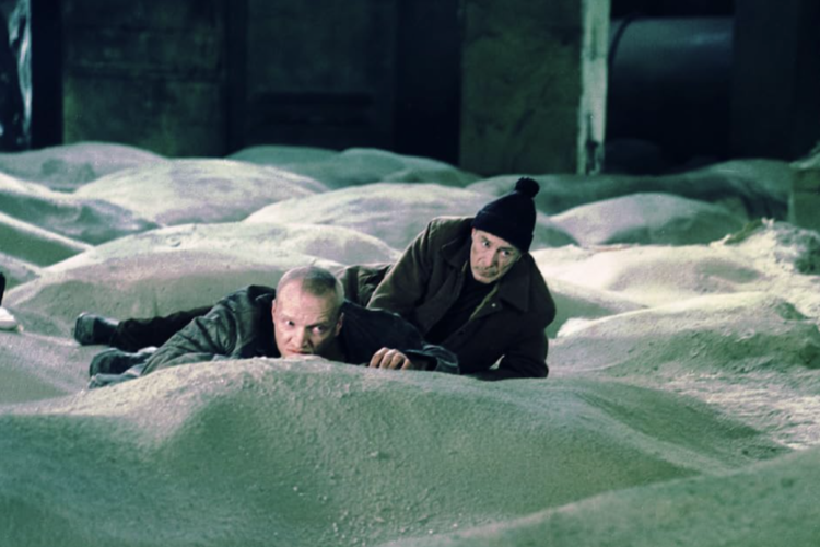Two men lie prone on a vast powdery substance, exchanging cautious glances in an industrial setting in this image from Mosfilm.