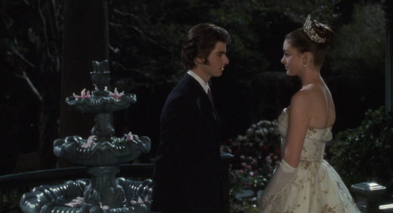 A man in a suit and a woman in a white and gold ball gown talk in a dark garden by a fountain in this image from Walt Disney Pictures. 