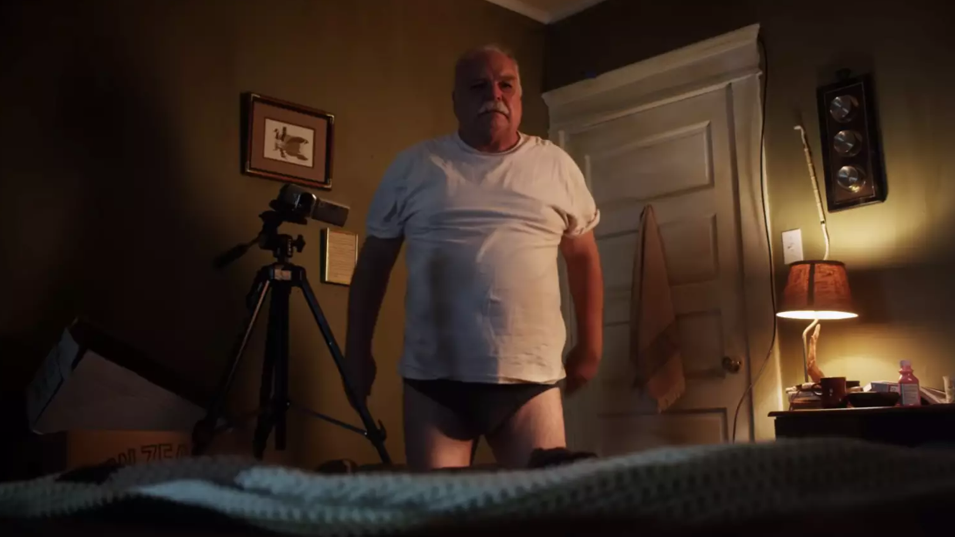 A man stands in a bedroom wearing a white T-shirt and underwear with a camera tripod next to him in this image from Independent.