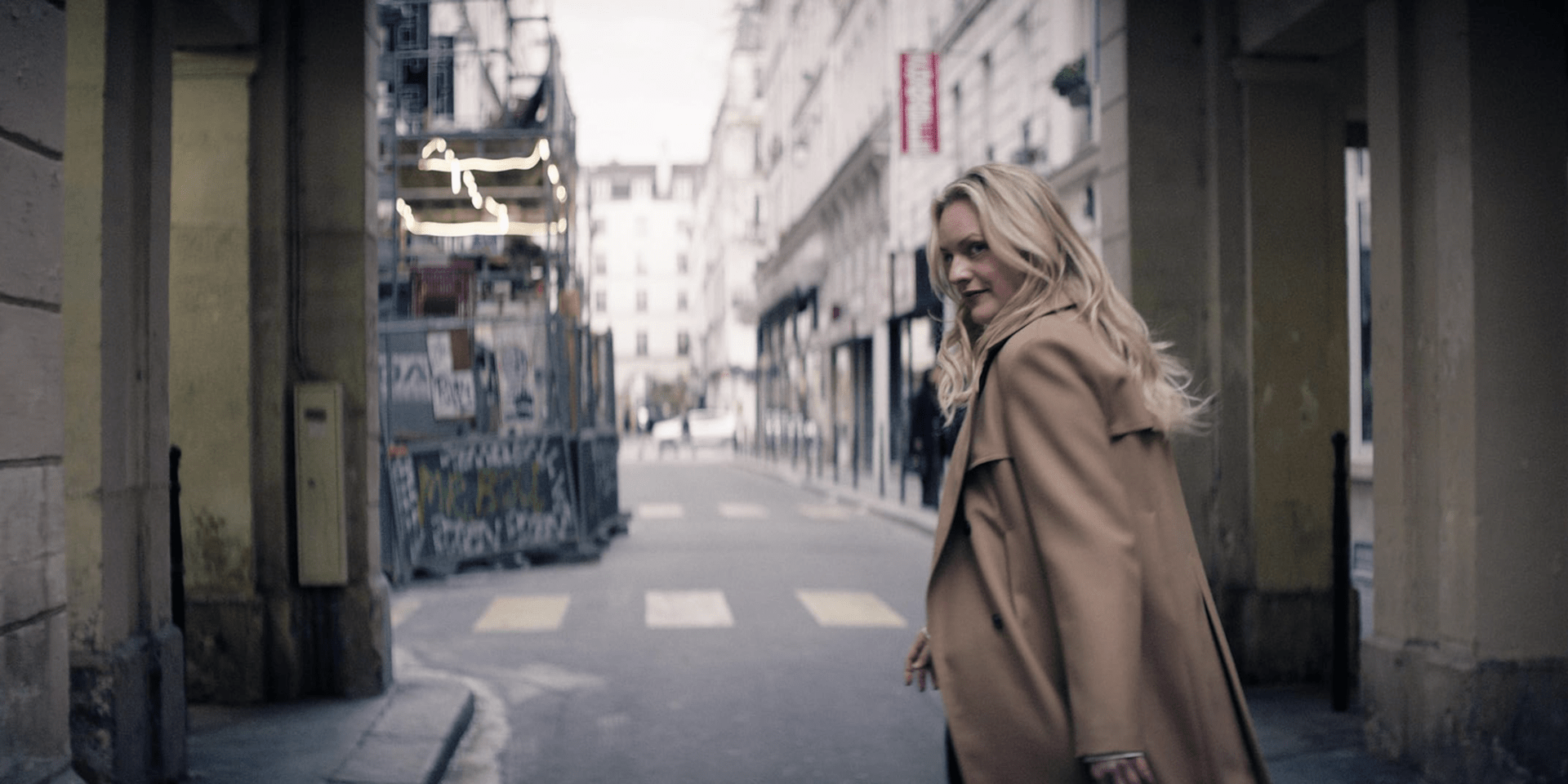 A woman in a trench coat looks over her shoulder while walking down an empty street in this image from Love & Squalor Pictures.